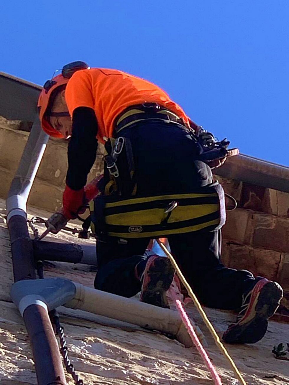 Cordiste intervenant sous une gouttière sur une façade en pierre à Saint-Paul-de-Vence.